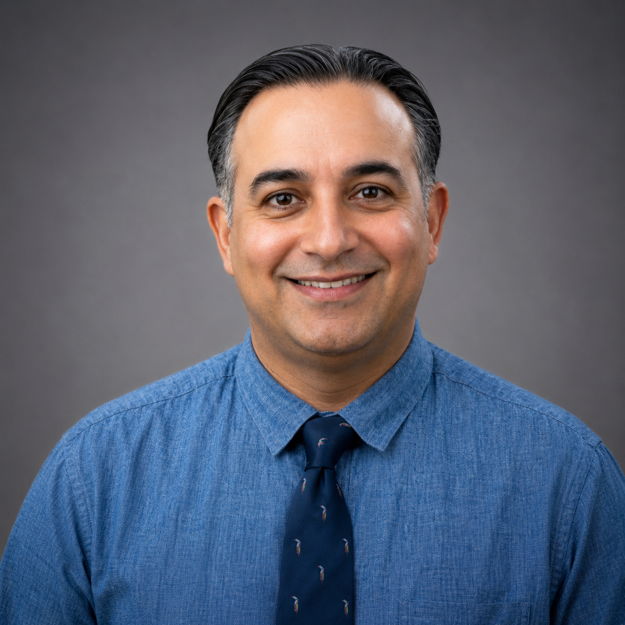 A man in a blue dress shirt and patterned tie smiling.